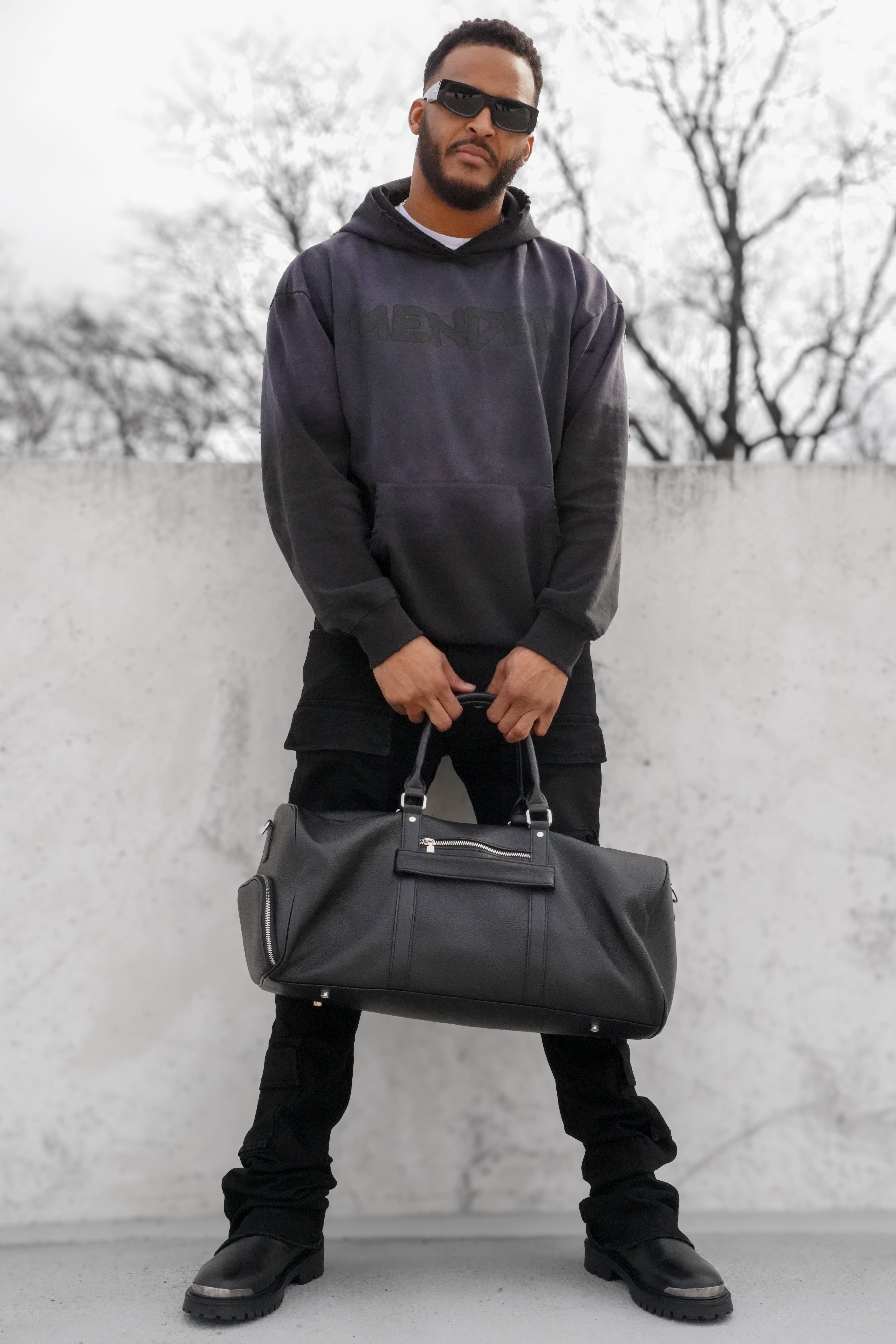 Man holding a black leather bag against a light-colored wall with trees in the background