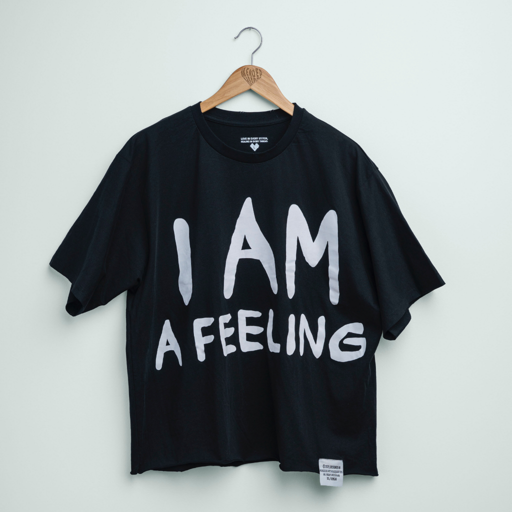 A black short-sleeve t-shirt hanging on a wooden hanger against a light gray wall. The shirt features the phrase 'I AM A FEELING' printed in large, white, hand-drawn style letters across the front.