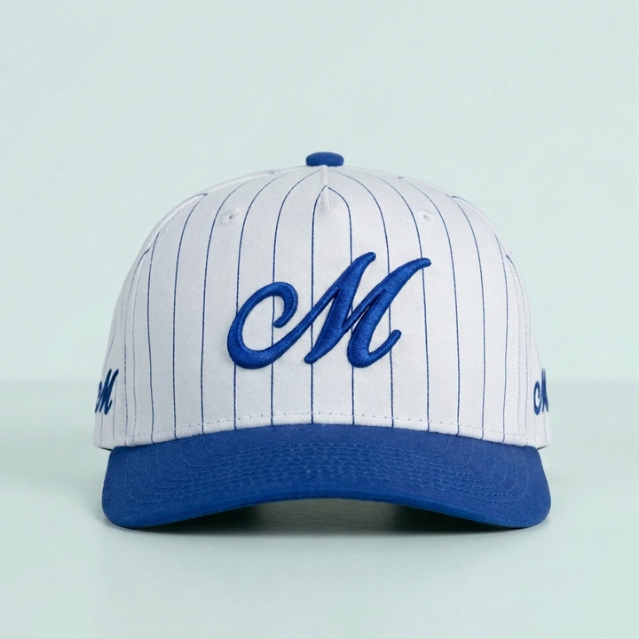 A baseball cap with a white crown featuring thin blue pinstripes, a royal blue visor, and a large embroidered blue cursive letter 'M' on the front. The cap is photographed against a solid pale mint green background.