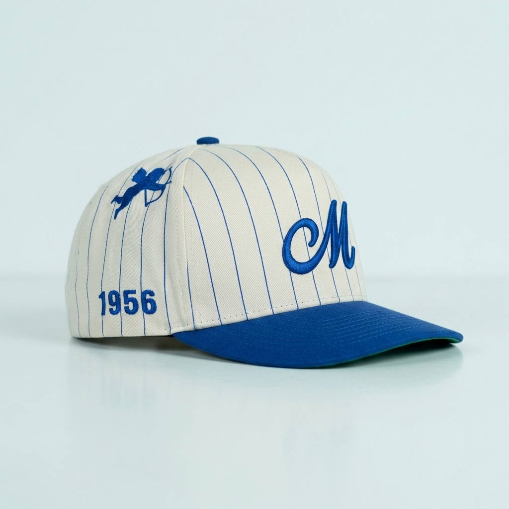 A baseball cap with an off-white pinstripe crown, a royal blue visor, and blue embroidered details including a large script 'M' on the front, a small winged emblem on the side, and '1956' on the other side.