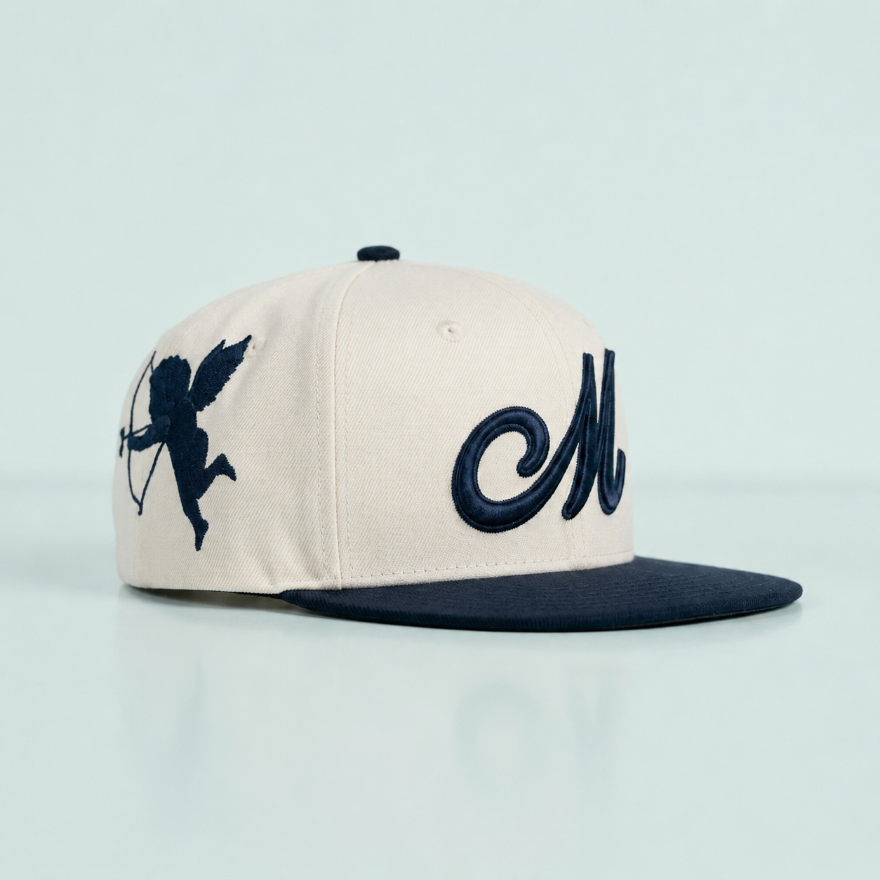 An off-white baseball cap with a navy blue flat brim, featuring a navy blue embroidered cupid silhouette on the side and a large cursive 'M' on the front, set against a light blue background.